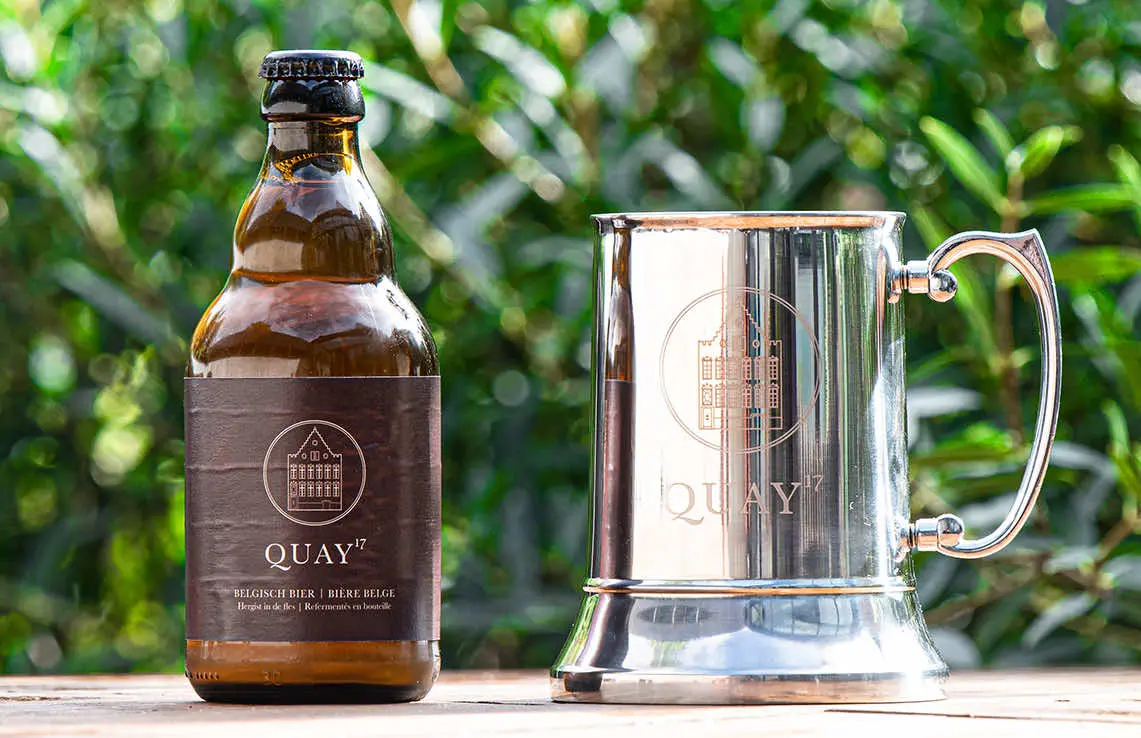 Fles Quay17-huisbier naast een zilveren bierkroes in het groen